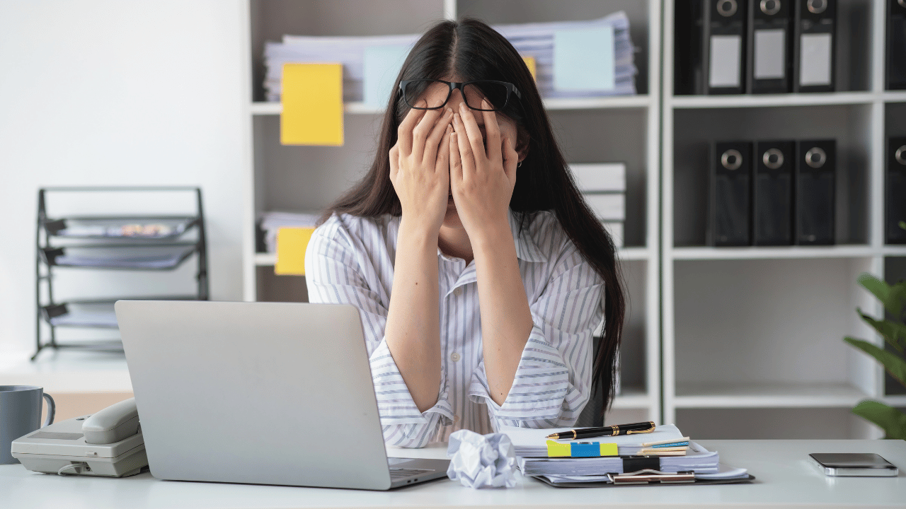 A dark-haired woman wearing a blue and white striped shirt sits at a desk with a silver laptop, clipboard and screwed up balls of paper. She is holding her head in her hands, pushing her glasses up to her forehead and obscuring her face. She emanates a sense of stress and frustration.